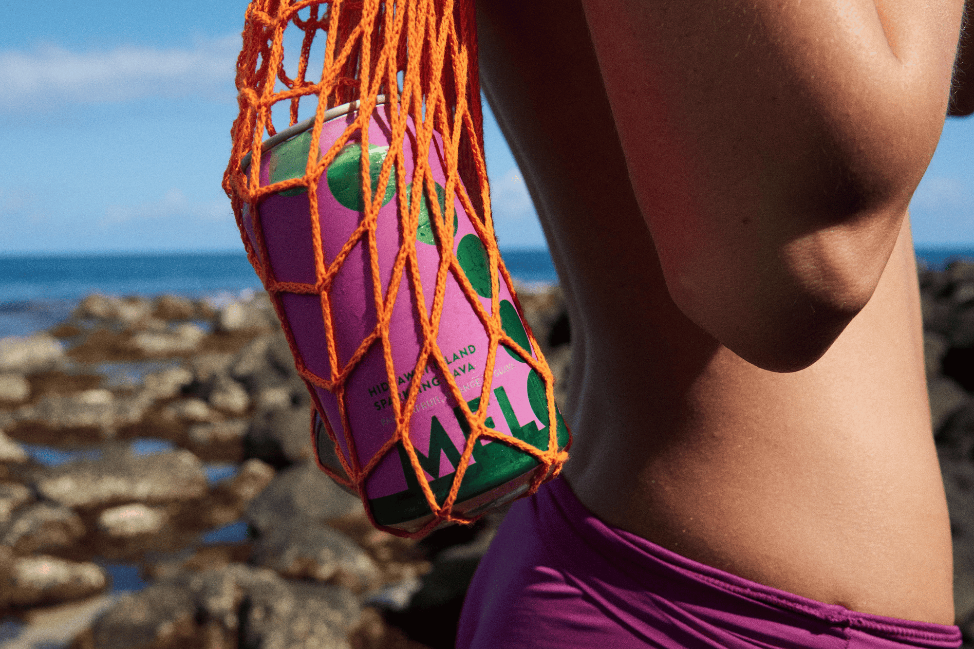 A pink Melo can in an orange net bag near a beach. Highlights Melo's tropical vibe and refreshing non-alcoholic beverages.
